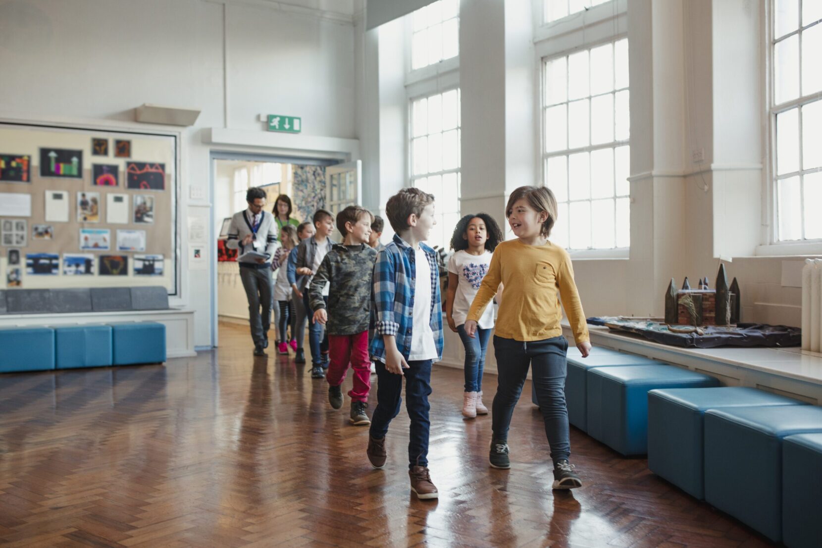 A group of elementary school children wearing casual clothing walk through the hall as their teachers walk behind them. This is a school in Hexham, Northumberland in north eastern England.